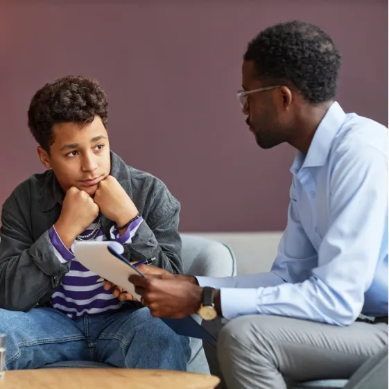 anxiety-teen-1 A teenage boy sits thoughtfully while talking to an adult man holding a clipboard.