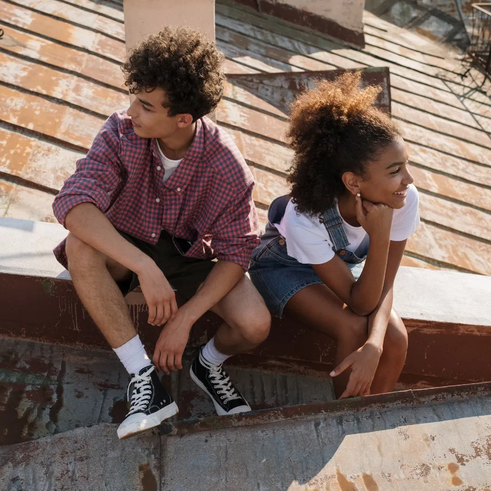Two teenagers sit on a slanted rooftop, smiling and enjoying the sunny weather.
