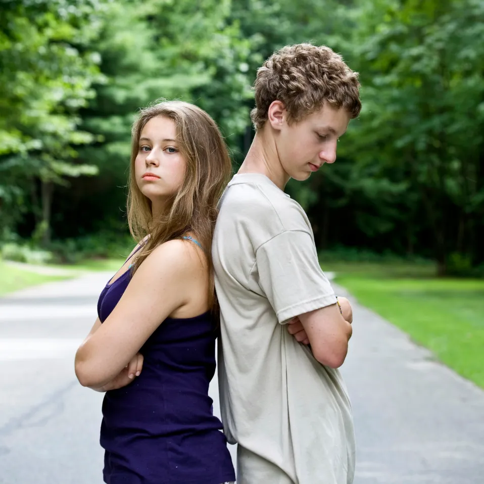 A teenage girl and boy stand back to back with arms crossed on a path in a green, wooded area.
