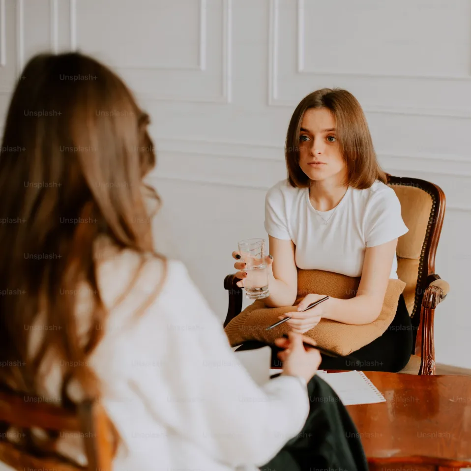 Woman holding a glass of water sits on a chair, talking to another woman in a therapy or counseling session.