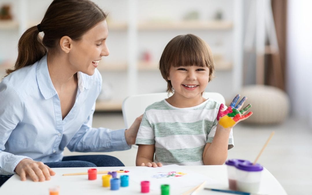 Smiling child shows painted hand while sitting with a woman at a table with art supplies.