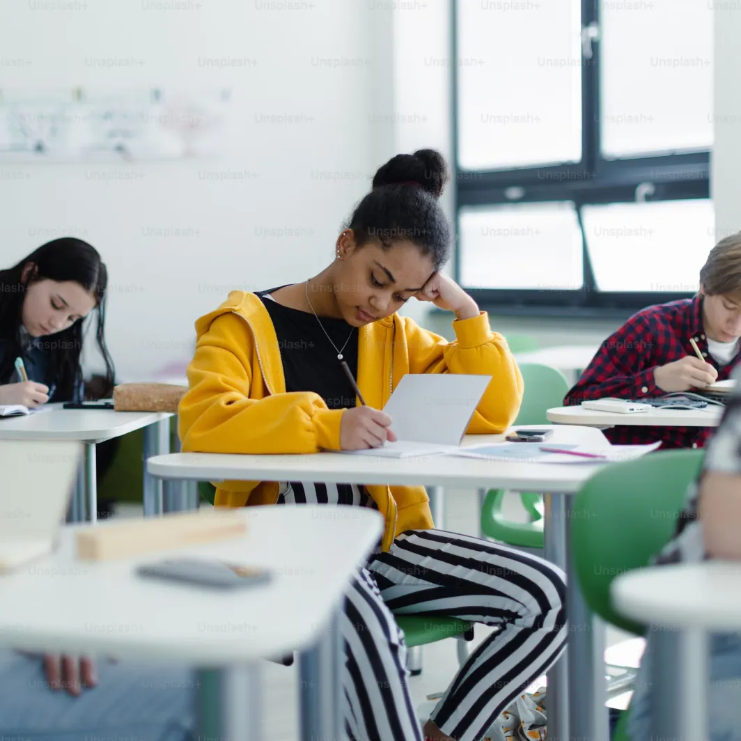 Student in a yellow hoodie writing at a desk in a classroom with others working in the background.