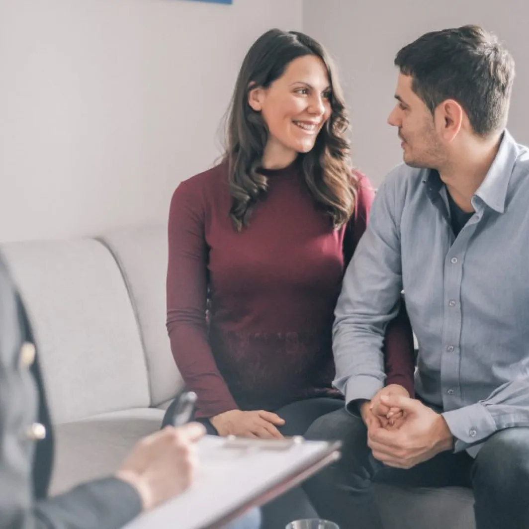 A smiling couple sits close together on a couch, talking with a therapist who is taking notes.
