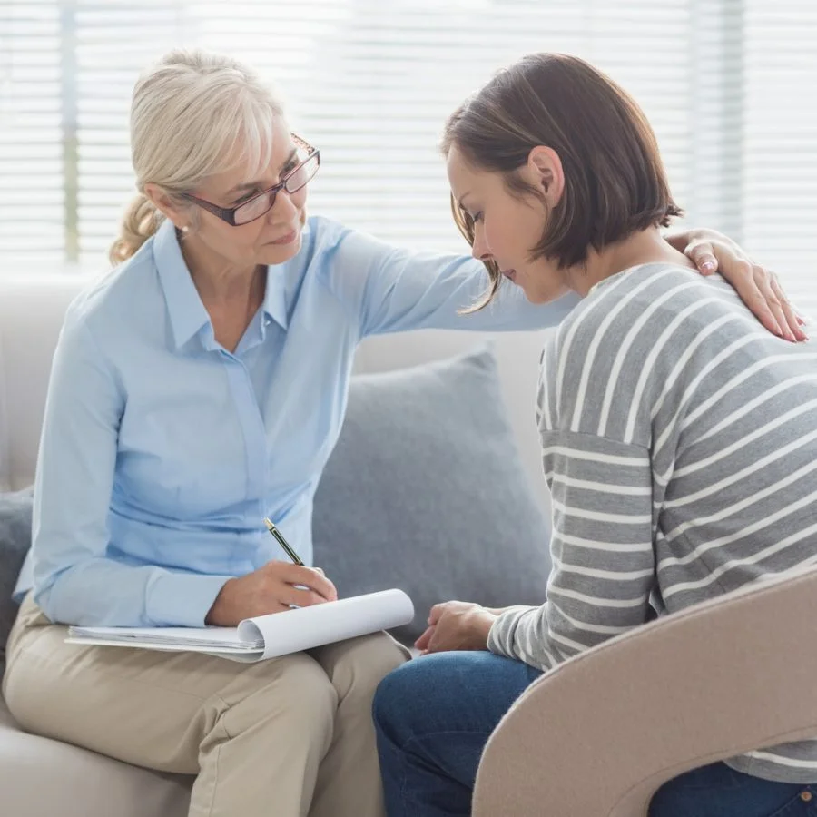 A therapist comforts a woman on a couch, gently touching her shoulder while taking notes.