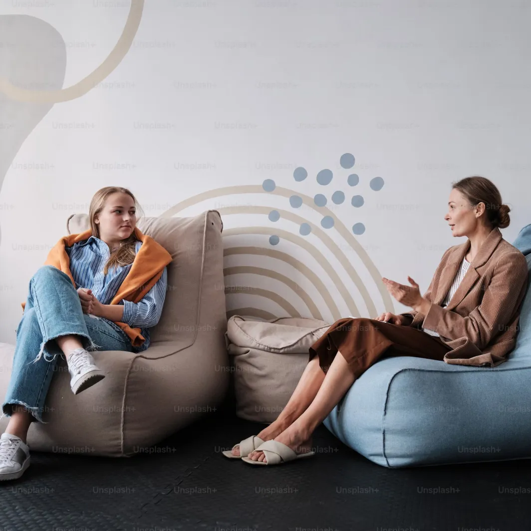 Two women sit on bean bag chairs, talking in a modern room with abstract wall art behind them.