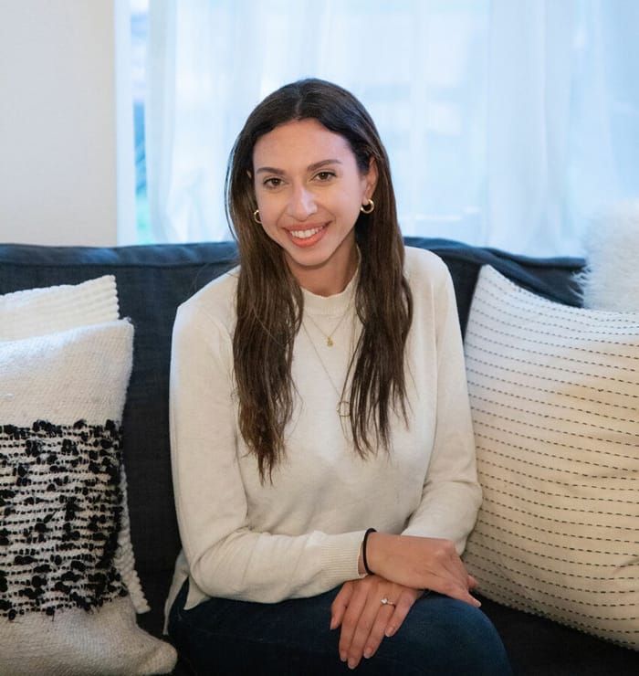 Woman with long brown hair smiles, sitting on a couch with white and patterned pillows in a cozy room.
