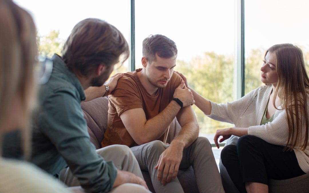 Three people comfort a sad man sitting on a chair in a bright room, offering support with gentle touches.
