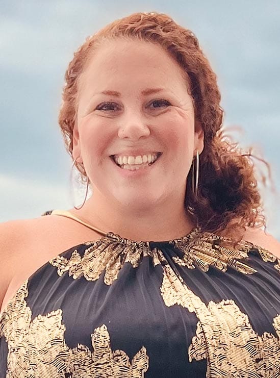 jennifer-cogan Smiling woman with curly red hair, wearing a black and gold dress, standing outdoors with a blue sky background.