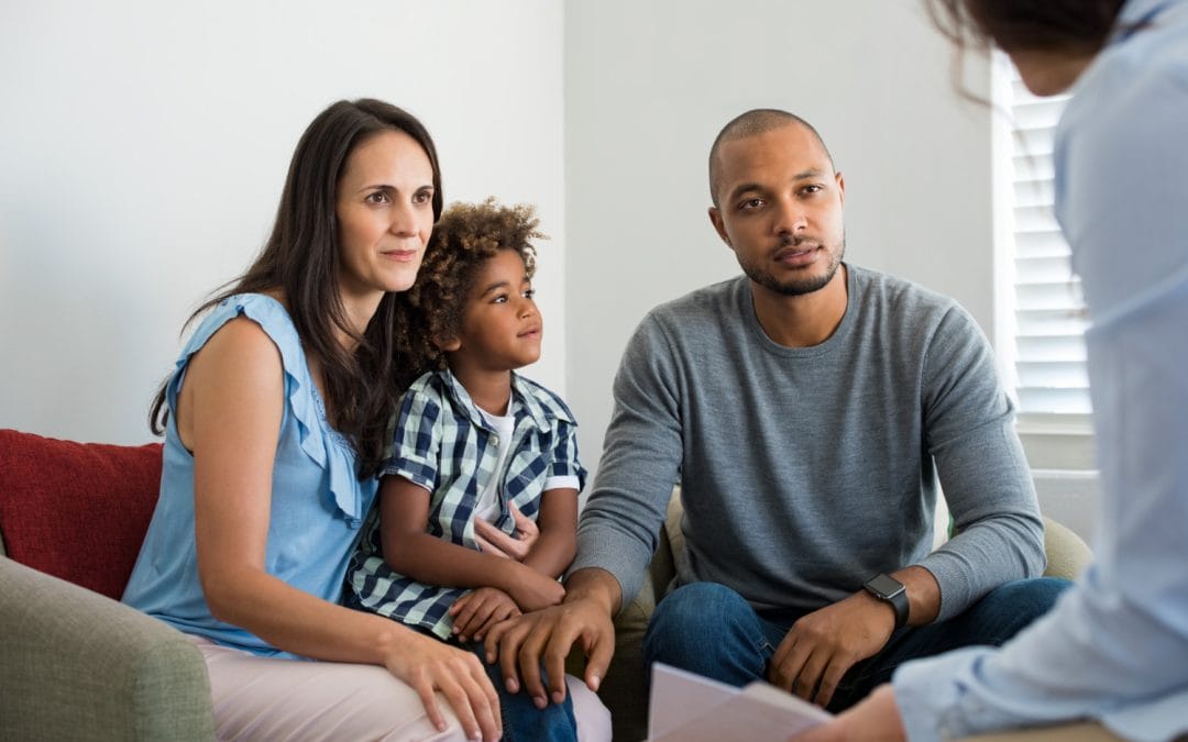 A family with a young child sits on a couch, attentively listening to a person holding a clipboard.