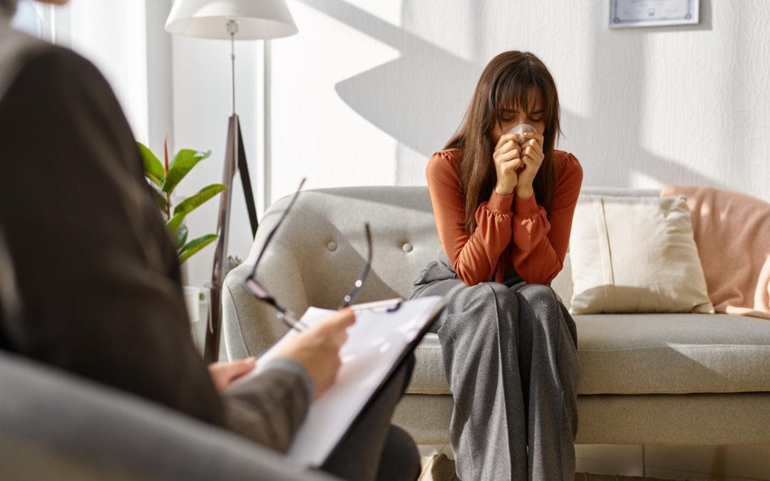 A woman sits on a couch crying while a therapist takes notes during a counseling session.