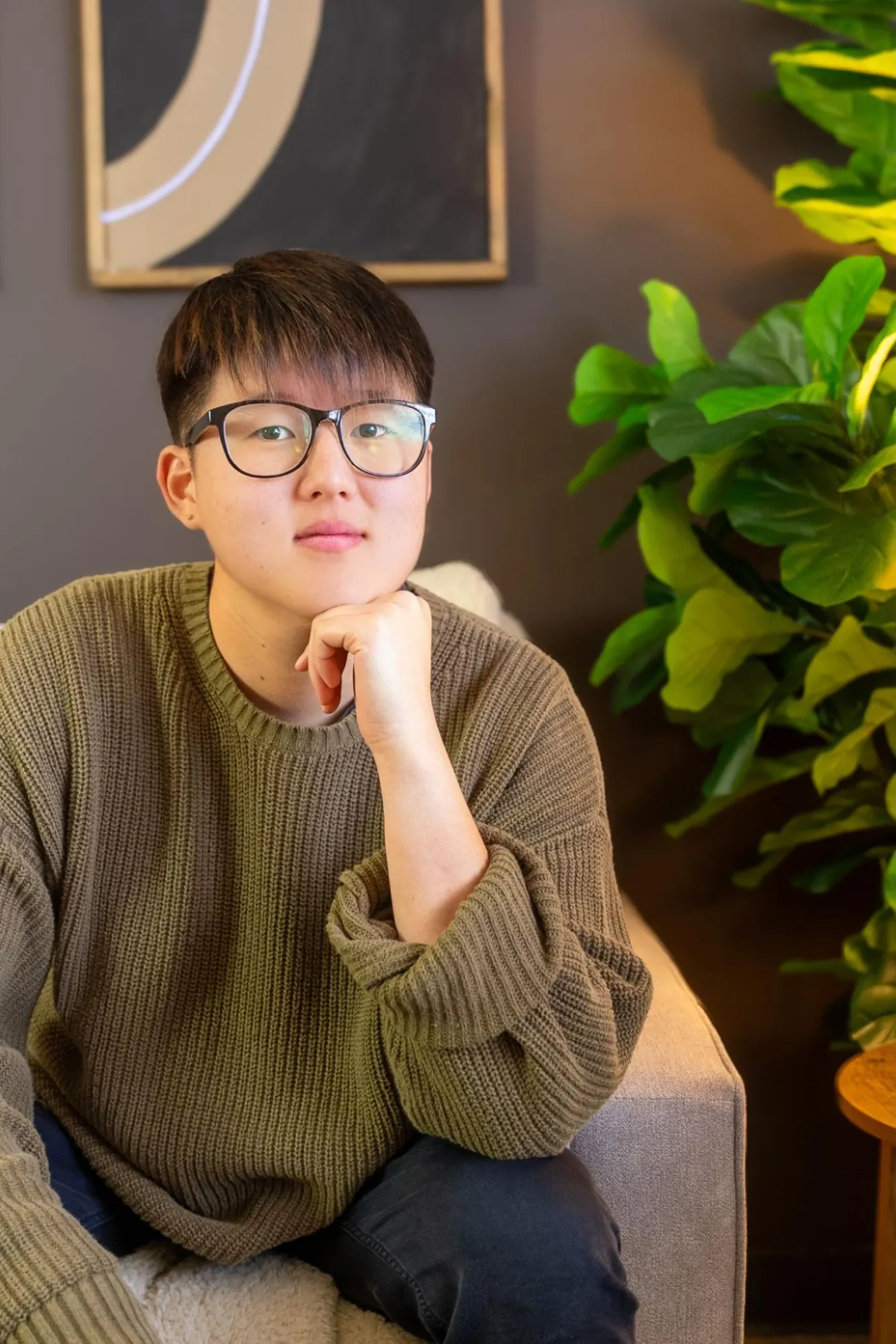 A person with short hair and glasses sits indoors on a couch, wearing a green sweater, next to a leafy plant.