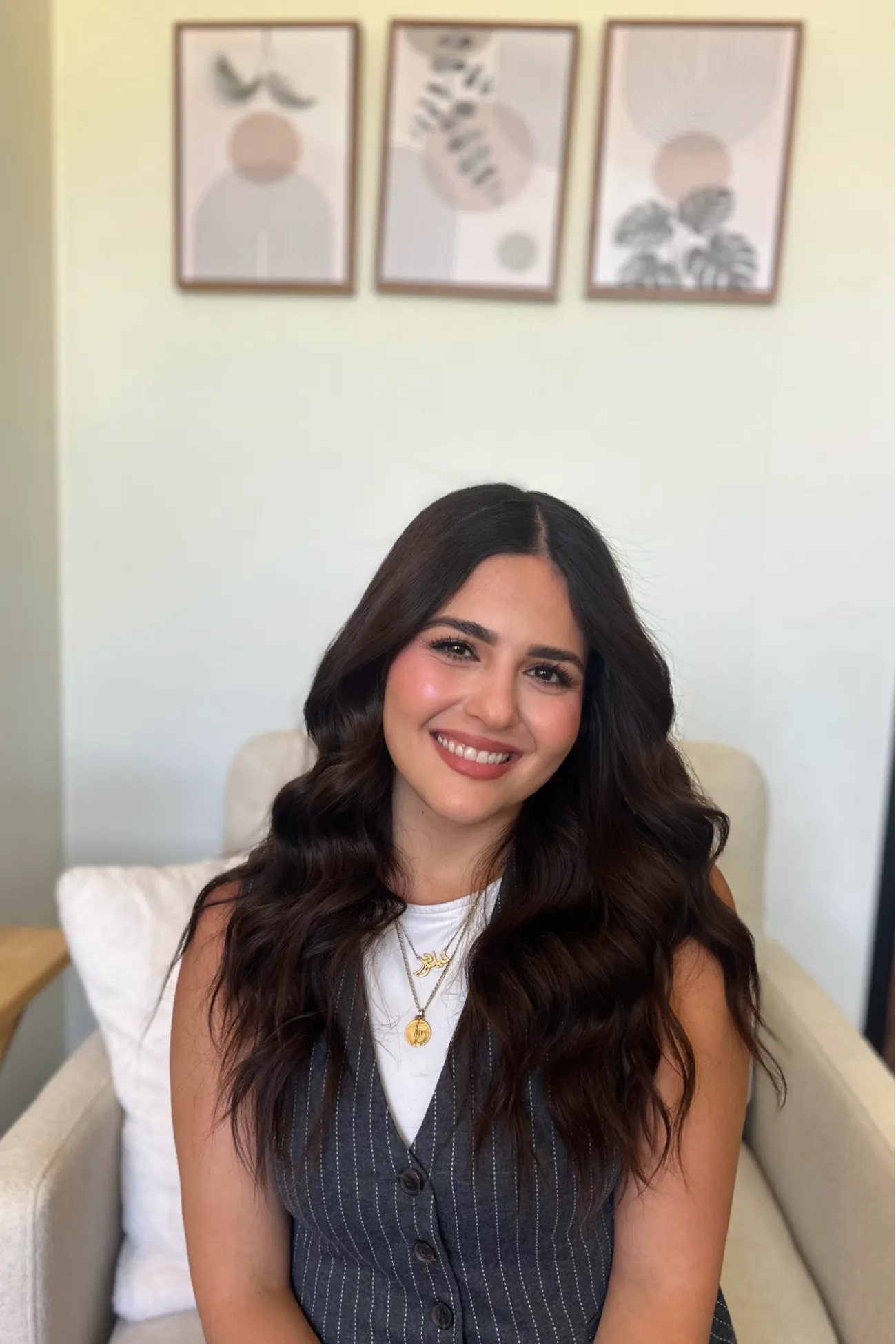 A woman with long wavy hair smiles while sitting on a chair, with three framed artworks on the wall behind her.