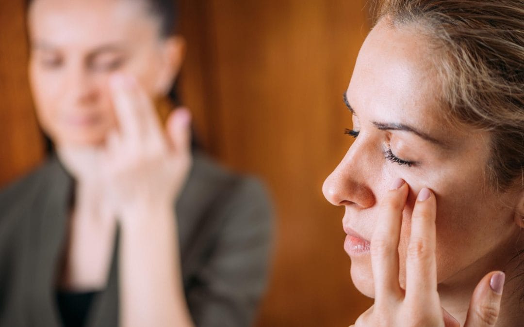Two women gently touching their faces near the eyes, focusing with closed eyes in a calm, indoor setting.