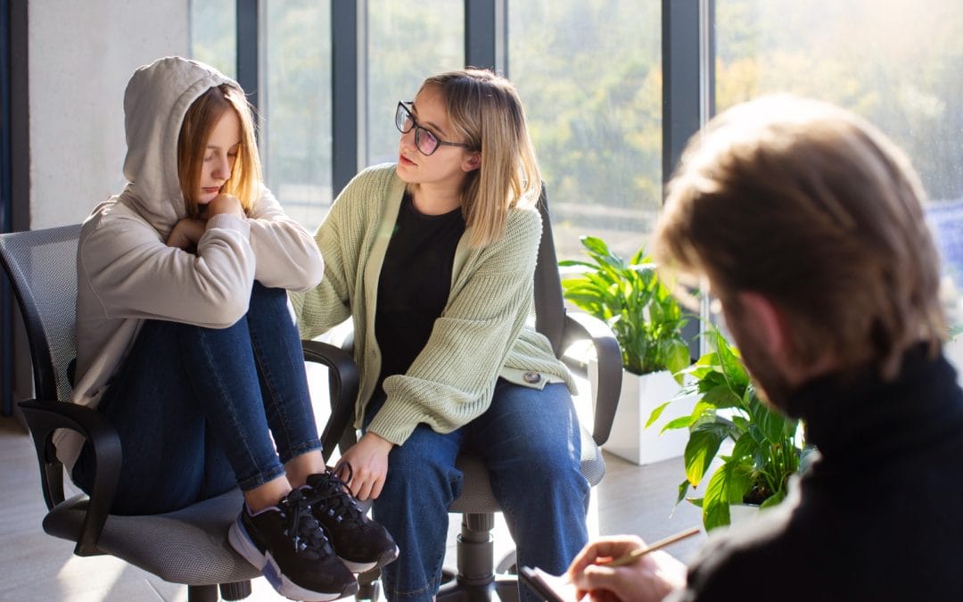 A girl sits with knees up, comforted by a woman, while a man takes notes in a bright room.