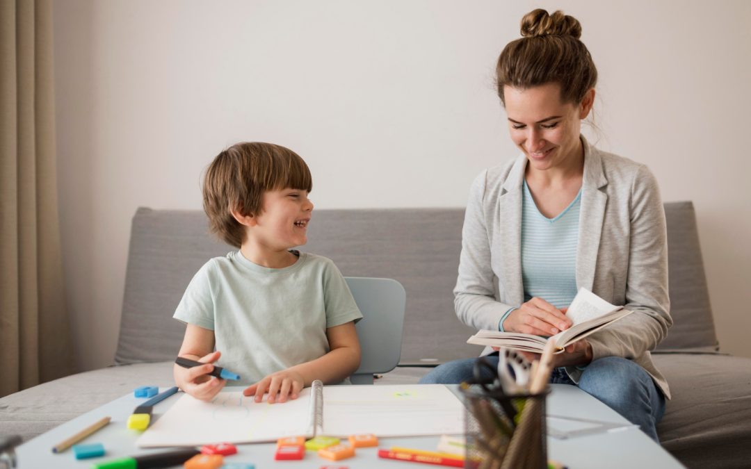 Smiling woman reads with a young boy as they do homework together at a table with school supplies.