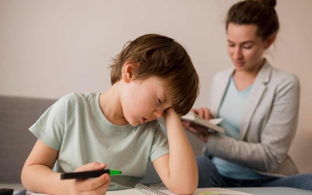 Young boy looking tired while drawing, with a woman reading in the background.