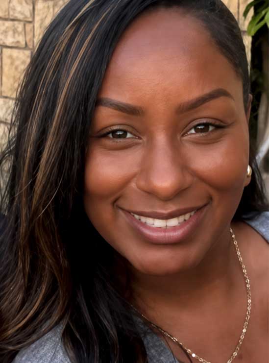 Smiling woman with long dark hair, wearing a gray top and gold necklace, standing outdoors by a stone wall.