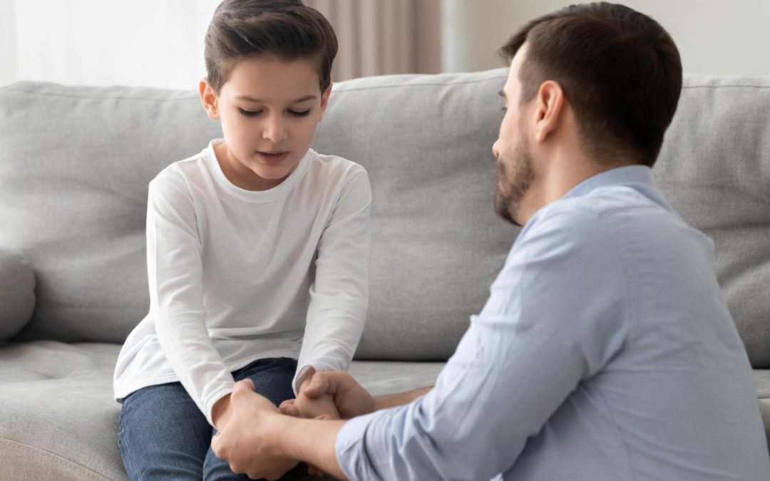 A man holds a boy’s hands, comforting him while they sit on a couch at home.
