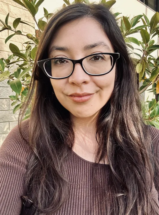 Woman with long dark hair and glasses, smiling outdoors in front of green leafy plants.