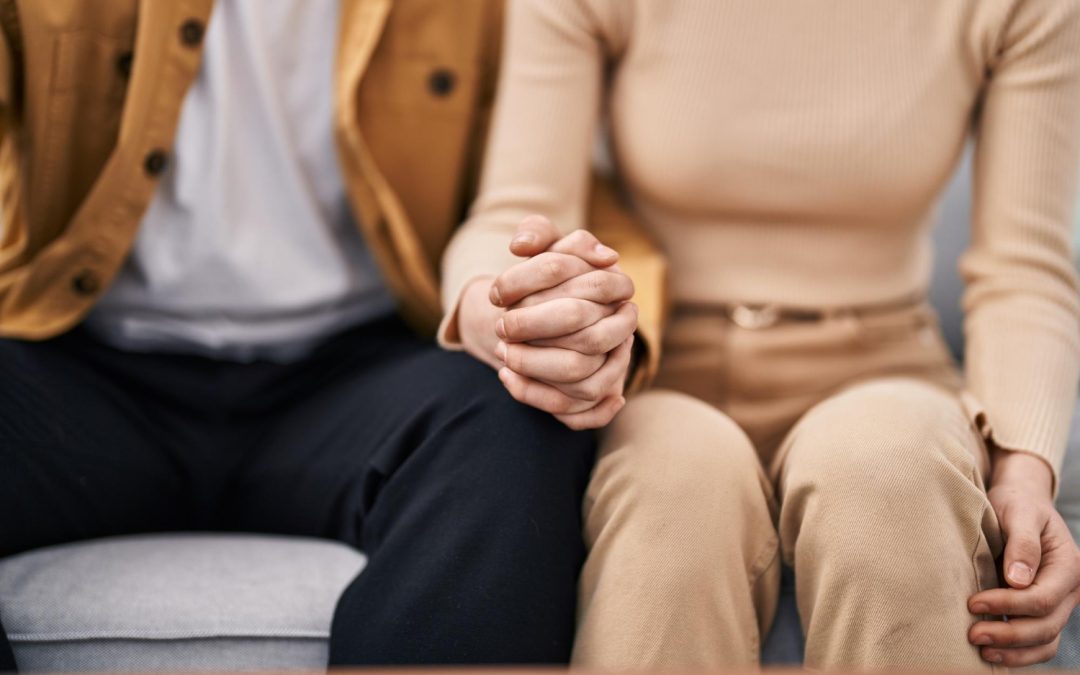 Two people sitting side by side on a couch, holding hands, only their torsos and legs visible.