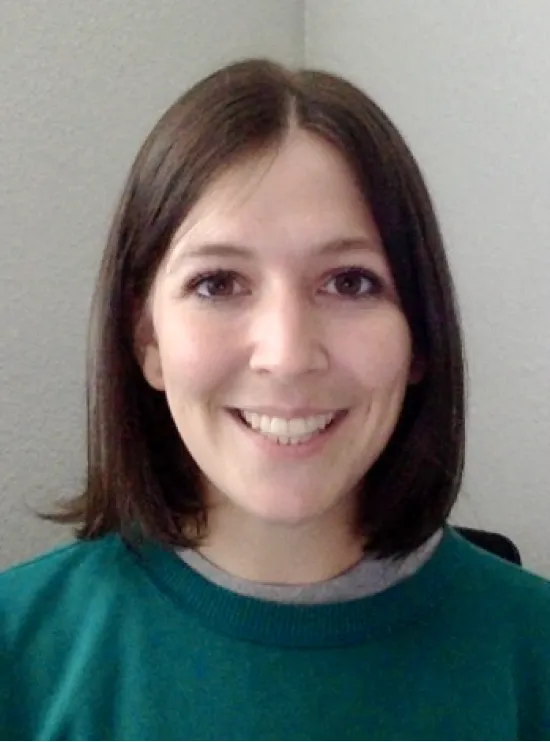 Woman with shoulder-length brown hair, wearing a green sweater, smiling at the camera against a gray wall.