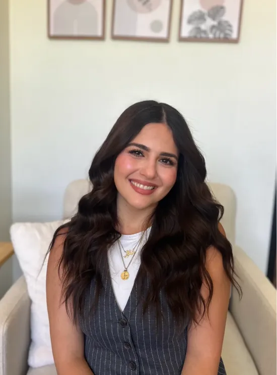 Woman with long wavy dark hair, sitting on a chair and smiling, wearing a vest and layered necklaces.
